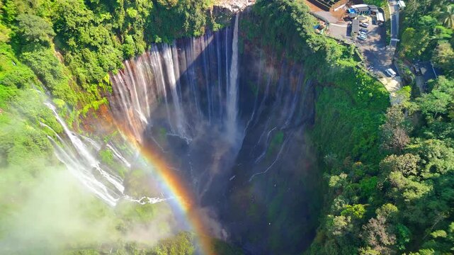 Aerial view of Tumpak Sewu Waterfall featuring a vibrant rainbow over lush green cliffs and mist in Kecamatan Pronojiwo, Jawa Timur, Indonesia.