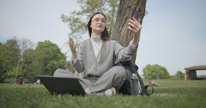 A hand-held shot of a young, beautiful female student in casual clothes, wearing glasses and headphones, sitting on the grass under a tree in a city park, using a laptop and talking on a mobile phone.