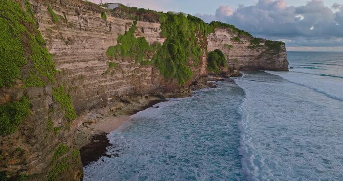 Bukit Peninsula tropical coast with high limestone cliffs covered in green vegetation rising above the ocean, waves crashing on a small sandy beach below during a colorful sunset. Aerial drone footage