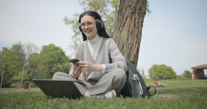 A hand-held shot of a young beautiful female student in casual clothes, wearing glasses and headphones, sitting on the grass under a tree in a city park and using a laptop while typing on the keyboard