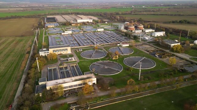 Aerial view of a wastewater treatment plant with multiple large circular clarifier tanks, sedimentation basins, channels and filtration structures arranged in a geometric industrial water-processing c
