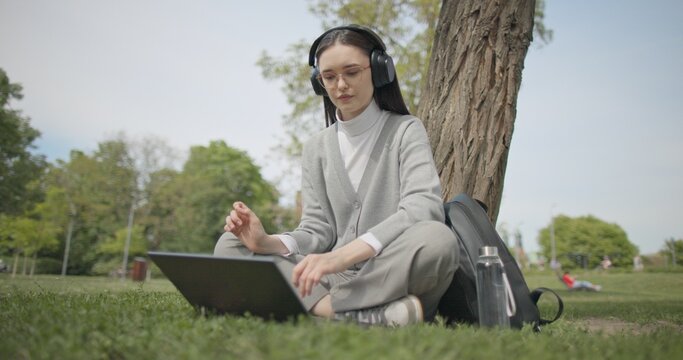 A hand-held shot of a young beautiful female student in casual clothes, wearing glasses and headphones, sitting on the grass under a tree in a city park and using a laptop while typing on the keyboard