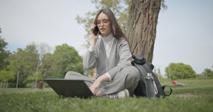 A hand-held shot of a young, beautiful female student in casual clothes, wearing glasses and headphones, sitting on the grass under a tree in a city park, using a laptop and talking on a mobile phone.