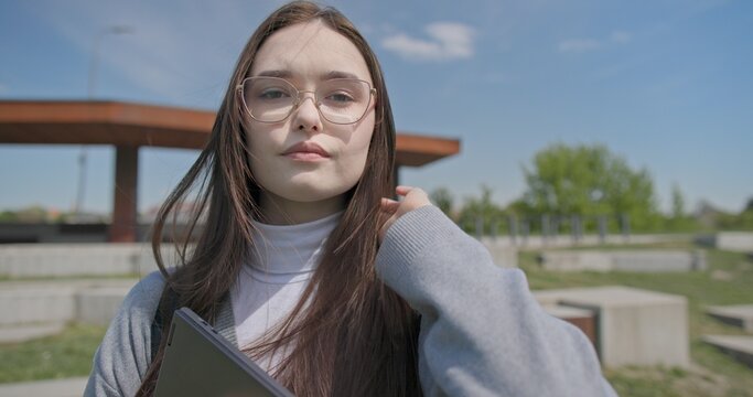 A hand-held portrait of a young female student in casual clothes, wearing glasses and a backpack, holding a notebook and laptop in her hands and posing looking at the camera while standing in a park 