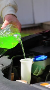 Close-up of hands pouring summer windshield washer fluid into reservoir under car hood, refilling green screen wash liquid in engine bay outdoors, car maintenance and care, vehicle service routine and