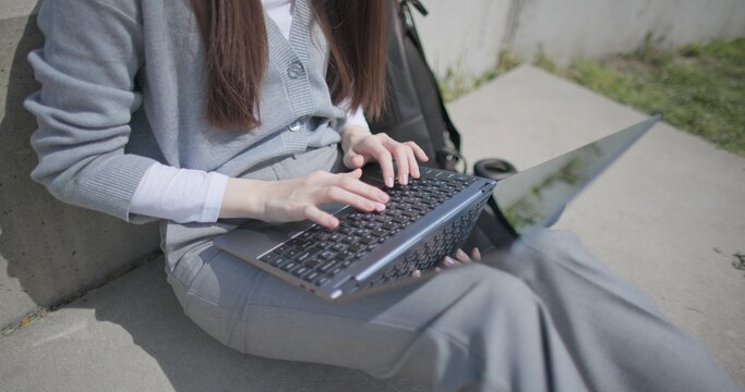 A hand-held shot of a young female student in casual clothes, glasses, and headphones working and typing on a laptop while sitting on a concrete bench in an urban park.