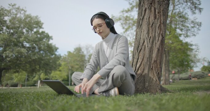A hand-held shot of a young beautiful female student in casual clothes, wearing glasses and headphones, sitting on the grass under a tree in a city park and using a laptop while typing on the keyboard