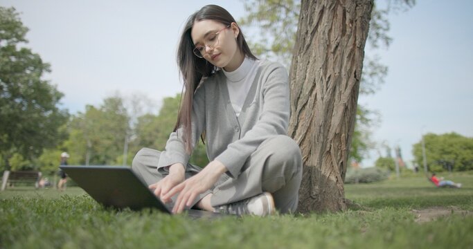 A hand-held shot of a young female student in casual clothes wearing glasses sitting on the grass under a tree in a city park and using a laptop typing on a keyboard.