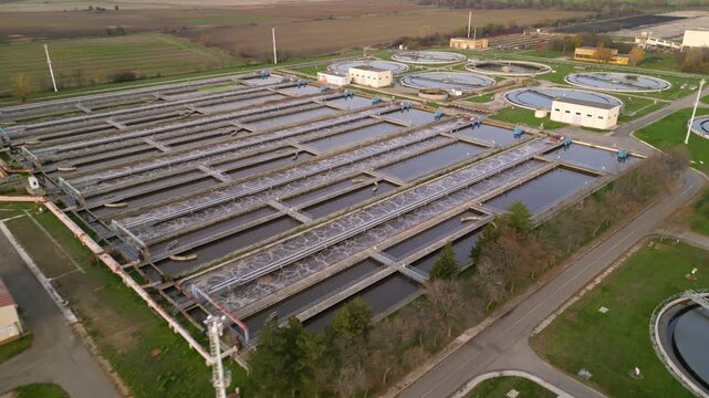 Aerial view of a large wastewater treatment plant with circular settling tanks, rectangular basins, pumping facilities, and green service roads set among agricultural fields and a canal under soft ove