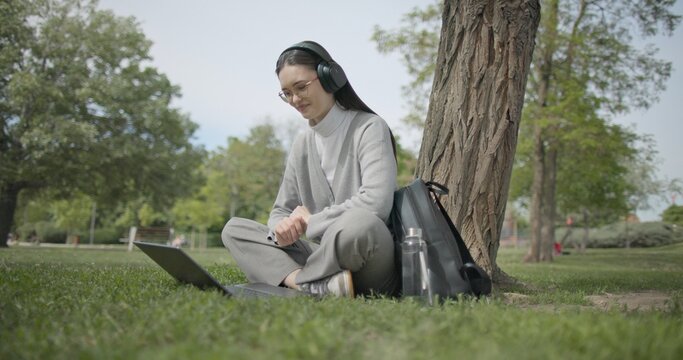 A hand-held shot of a young beautiful female student in casual clothes, wearing glasses and headphones, sitting on the grass under a tree in a city park and using a laptop while typing on the keyboard
