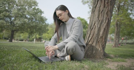 A hand-held shot of a young female student in casual clothes wearing glasses sitting on the grass under a tree in a city park and using a laptop typing on a keyboard. © Anatoly Repin