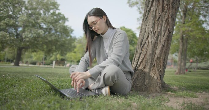 A hand-held shot of a young female student in casual clothes wearing glasses sitting on the grass under a tree in a city park and using a laptop typing on a keyboard.