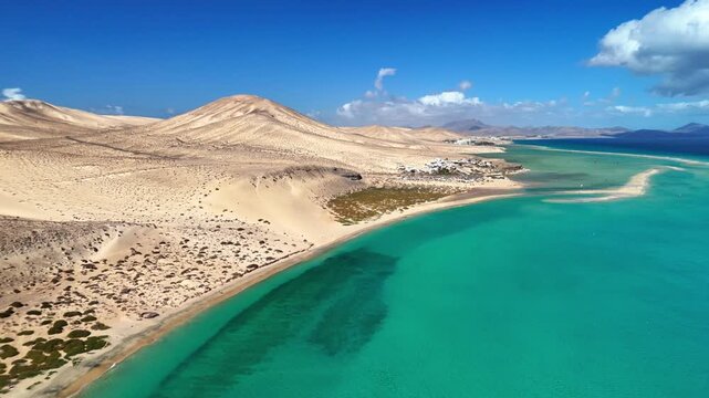 Aerial view of Playa del Matorral featuring turquoise waters, vast sand dunes, and volcanic hills under a clear blue sky in Canary Islands, Spain.