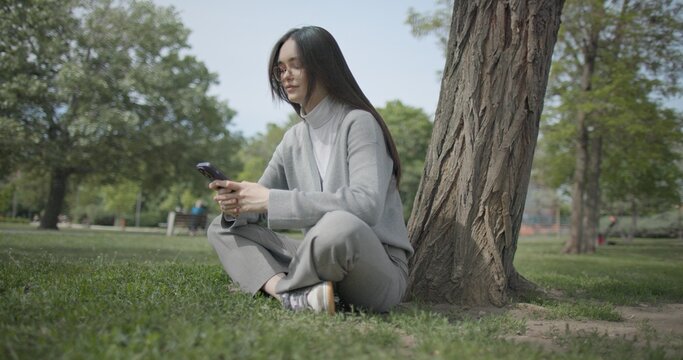 The shot is handheld and shows a young female student in casual clothes wearing glasses sitting on the grass under a tree in a park and using a mobile phone.