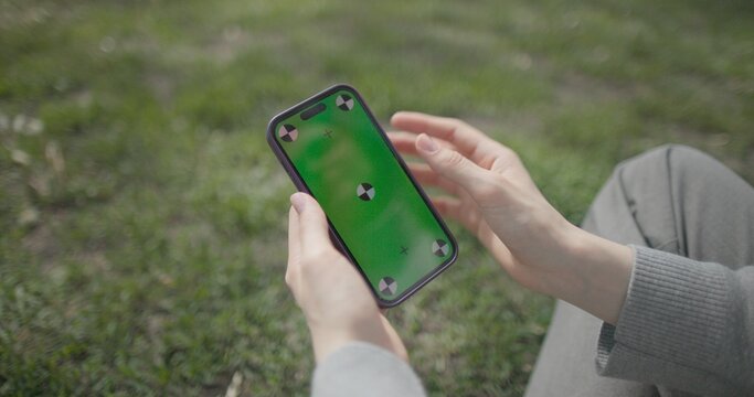 A close-up shot of female hands holding a mobile phone against a background of grass and making gestures of using the phone