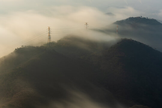 Amazing sea of clouds flowing like a waterfall over a mountain ridge with electricity pylons during a beautiful pastel sunrise.