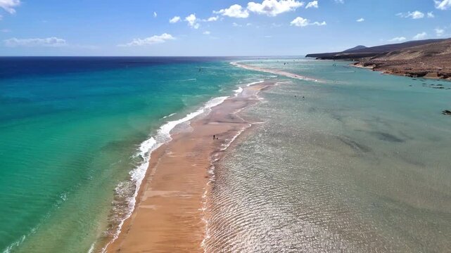 Aerial view of Playa del Matorral featuring a long sandbar, turquoise ocean waves, and volcanic hills under a blue sky with scattered clouds, Canary Islands, Spain.