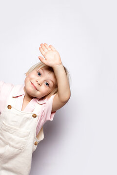Cute blonde toddler boy waving hand or saying hi. Happy friendly little child looking at camera with head tilt. Vertical studio portrait of sweet kid in overalls pink shirt on white background