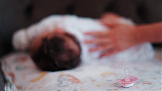 Newborn baby sleeping peacefully in bed with mother hand gently patting infant back in cozy home nursery setting.