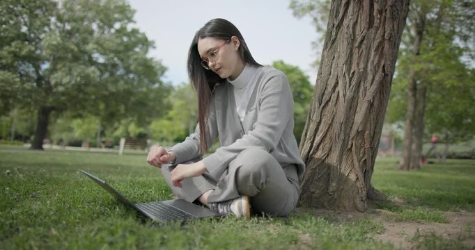 A hand-held shot of a young female student in casual clothes wearing glasses sitting on the grass under a tree in a city park and using a laptop typing on a keyboard.