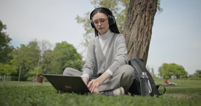 A hand-held shot of a young beautiful female student in casual clothes, wearing glasses and headphones, sitting on the grass under a tree in a city park and using a laptop while typing on the keyboard