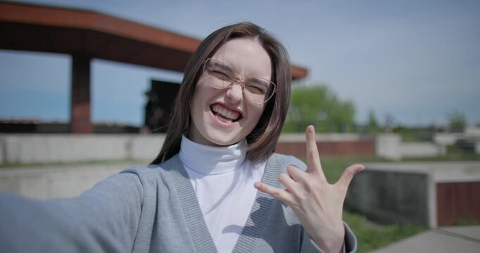The shot is handheld and shows a young female student in casual clothes wearing glasses, sitting and recording a selfie video on her phone, holding it in her hand and gesturing with her other hand.