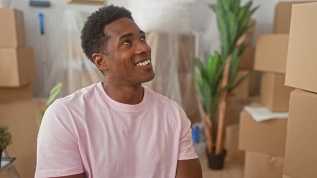 Man smiling and looking up amid stacked moving cardboard boxes in building, wearing a pink t shirt and relaxed posture; joy accomplishment.