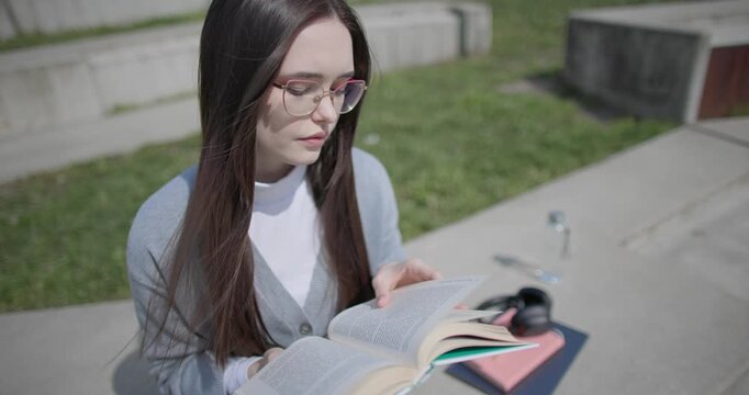 A hand-held shot of a young female student in casual clothes wearing glasses sitting on a concrete bench in an urban park on a sunny summer day and reading a book, flipping through the pages.