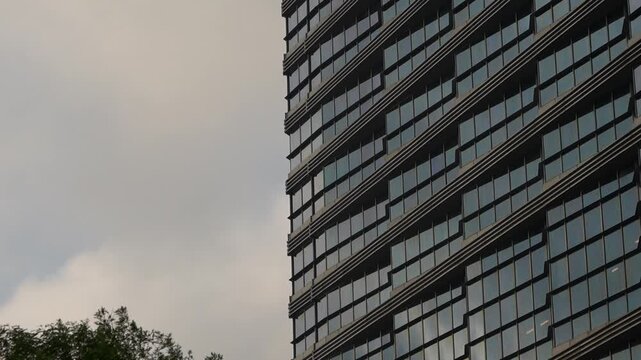 This is a partial close-up shot of a modern high-rise building's facade, with glass curtain walls and staggered balcony structures, set against a cloudy overcast sky with treetops at the bottom left.