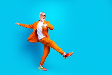 Obraz premium Joyful senior man dancing in bright orange suit against a blue background, stylish retiree celebrating life with energetic pose and colorful sneakers