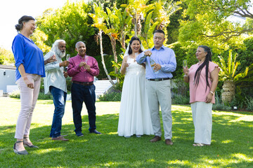 Groom popping champagne, bride wearing wedding gown holding flute, guests celebrating on lawn © wavebreak3