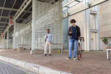 Fototapeta premium Two adult males waiting at curbside, one in leather jacket with bicycle checking phone, copy space