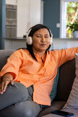 Fototapeta premium Mature Asian woman sitting on gray sofa at home, listening on white headphones, smartphone beside
