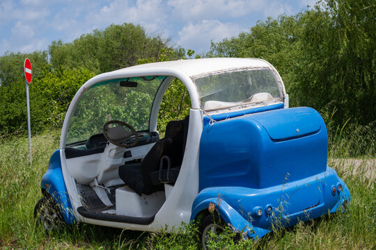 A small, doorless blue and white electric utility cart sits abandoned in overgrown grass near a "no entry" traffic sign on a sunny day