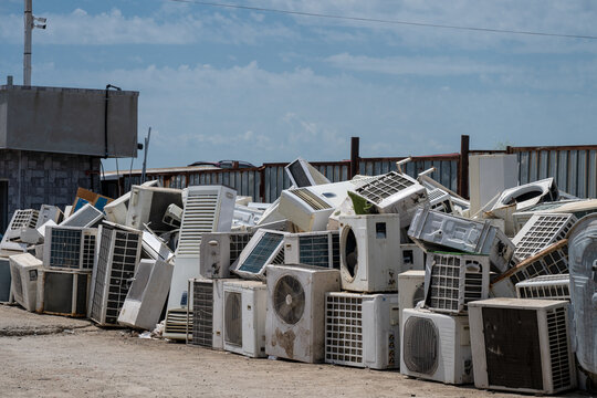 A massive pile of discarded and broken air conditioner compressor units sits outdoors in a junkyard under a blue sky.