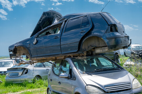 A stripped dark blue hatchback missing its wheels sits stacked precariously on top of a silver car in a grassy, outdoor auto salvage yard.