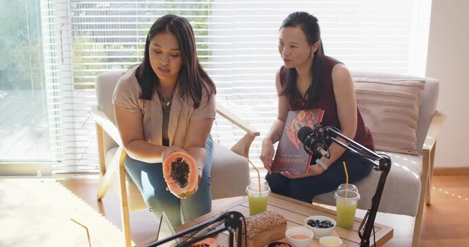 Diverse female friends showing halved papaya in living room, left tilting to laptop mic for demo
