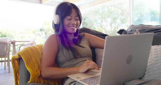 Settling on couch, Asian woman typing on silver laptop wearing white headphones while working