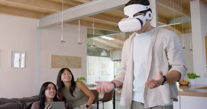 Three Asian friends watching man putting on white VR headset and showing virtual objects at home
