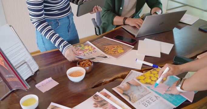 Diverse team using tablet pointing prints marking sorting covers at meeting table reviewing designs