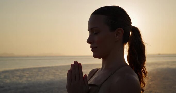 Portrait of happy woman prays holding hands in namaste mudra on sunset sea beach. Contemplative lady engages in meditation and mindfulness practices at seaside