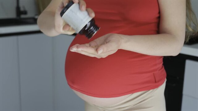 Pregnant woman pouring vitamin pills from bottle into palm in kitchen.