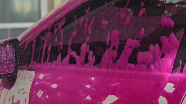 Pink drops of foam, car wash chemicals, drip down the side window of the car, contrasting sharply with the interior