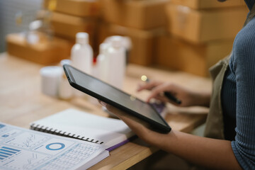 Female ecommerce entrepreneur using tablet to manage online business data and skincare product orders in small business workspace. © Kieng