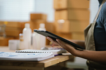 Female ecommerce entrepreneur using tablet to manage online business data and skincare product orders in small business workspace. © Kieng