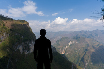 Silhouette stands atop cliff overlooking vast mountain range. Distant peaks stretch under bright, partly cloudy sky. Rugged terrain, lush green slopes, deep ravines visible below © jinhongljh