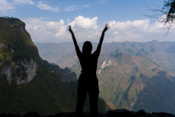 Silhouette stands with arms raised high. Mountain ranges stretch beneath a clear blue sky. Clouds drift across the distant horizon. Person embraces nature's vastness. Lush green valleys rise below © jinhongljh