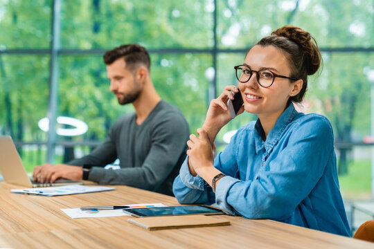 Two young attractive colleagues working together at an office desk, using a phone and laptop to collaborate on tasks, creating a dynamic and professional work environment.