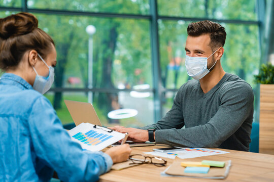 Two business partners sitting across from each other, wearing medical masks in a spacious office, discussing plans and collaborating while maintaining safety protocols in a professional setting.