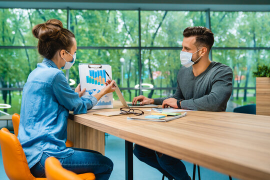 Two business partners discussing and showing diagrams at the office desk while wearing medical masks, collaborating on a project in a safe and professional work environment.